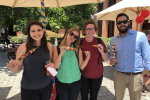 Four students stand together holding food kabobs at a lunch outside on a college campus.